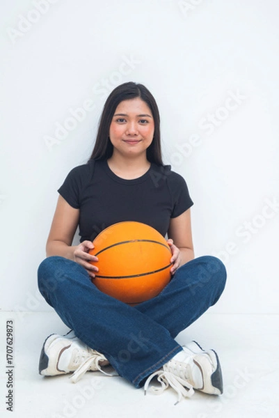 Fototapeta A bubbly young Asian woman in a half body shot sitting on the floor with legs crossed, smiling and holding a basketball with a focused and joyful demeanor. Isolated on a white background.