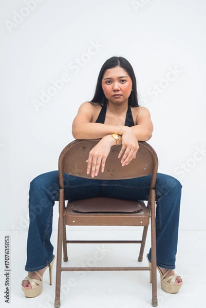 Fototapeta A fierce young Asian woman sitting on a backward chair with a strong, intense gaze that exudes confidence and power. Isolated on a white background.
