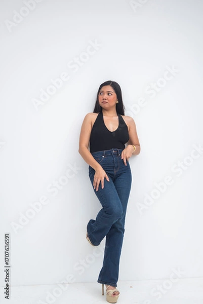 Fototapeta A graceful young Asian woman in a half body shot leaning on a wall, one foot resting on it while looking sideways with poise and sophistication. Isolated on a white background.