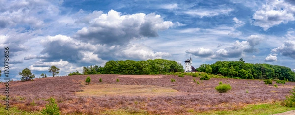 Fototapeta A scenic view of a country house with extraordinary architecture, surrounded by greenery atop a hill. Fluffy white clouds scatter across the vibrant blue sky, enhancing the natural beauty.