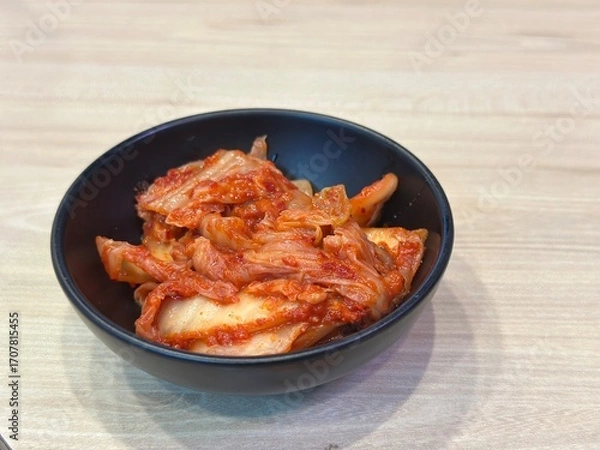 Fototapeta Authentic Korean Horizontal Shot: Spicy Fermented Napa Cabbage Kimchi (Baechu Kimchi) Served as a Side Dish in a Matte Black Bowl on a Light Wooden Table.