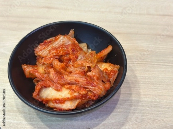 Fototapeta Authentic Korean Side Dish: Two Horizontal Shots of Spicy Fermented Napa Cabbage Kimchi (Baechu Kimchi) Served Generously in a Dark Matte Bowl on a Wooden Table.
