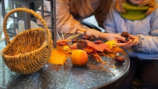 Obraz People engaging in creating autumn crafts using fallen leaves, chestnuts, and other natural materials on a table. A wicker basket and lantern add to the cozy seasonal ambiance.