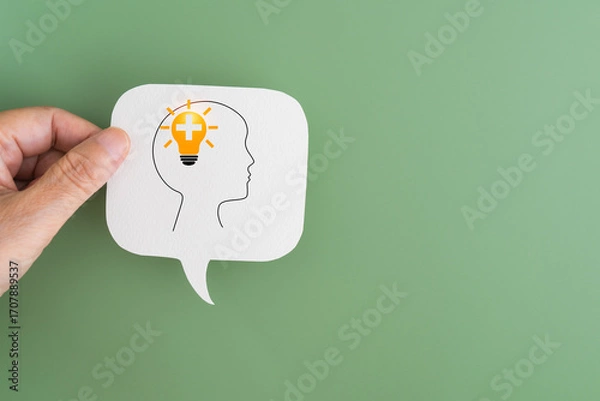 Fototapeta For positive thinking concept hand holding real white speech bubble paper cut with head and lightbulb with plus inside on green background