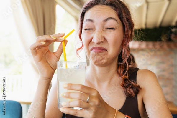Fototapeta Young woman making a disgusted funny face while drinking a glass of sour lemonade with a straw in a restaurant
