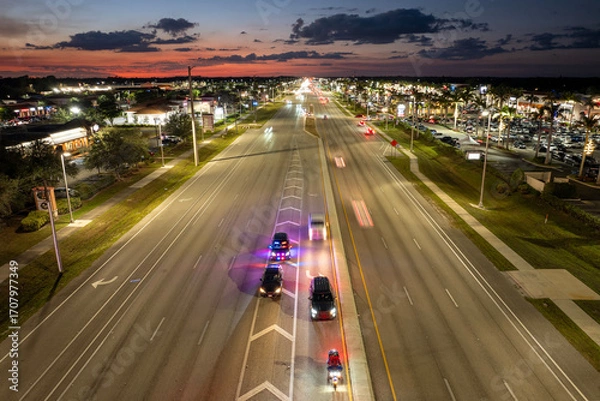 Fototapeta Police vehicle with emergency lights stops a driver for speeding on city road at night in the United States. Traffic moving rapidly on wide highway street.