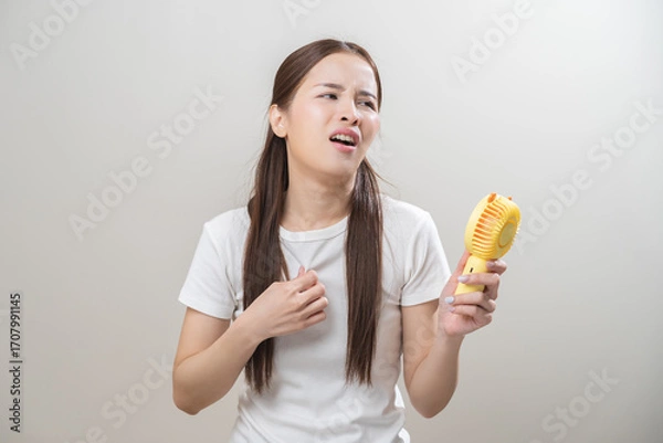 Fototapeta Suffering summer heat wave stroke, hot weather, tired asian young woman sweaty and thirsty, refreshing with hand in blowing, wave fan to ventilation when temperature high, isolated on background.