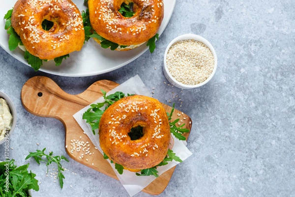 Fototapeta Three freshly baked bagels filled with greens and ham, on a wooden board and plates. Bagel breakfast sandwiches a zesty unique twist. top view