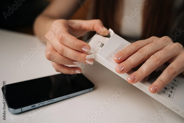 Fototapeta Close-up hands of happy woman with manicured nails carefully opening white retail package containing new phone adapter cable, ready to connect and charge. Concept of smartphone unboxing process.