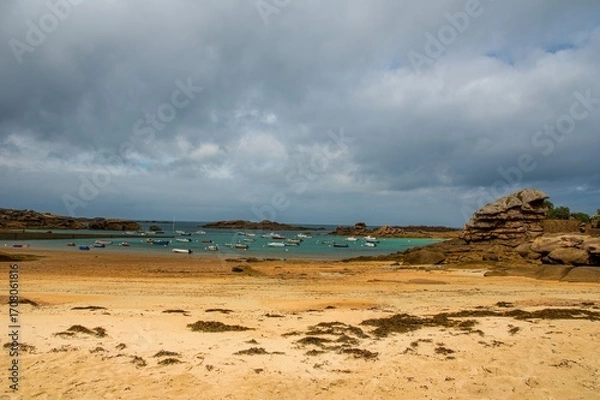 Fototapeta Sandy beach and turquoise bay at low tide with anchored boats and dramatic pink granite rock formations in Brittany, France, under a cloudy summer sky.