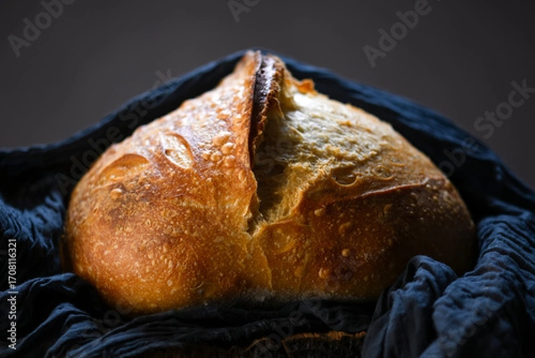 Fototapeta Appetizing sourdough bread with cracked crust and rugged texture wrapped in blue linen towel. Artisan farmhouse-style loaf captured in warm natural lighting