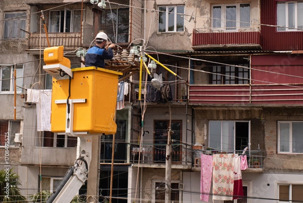Fototapeta An electrician in a hard hat in a yellow aerial work platform repairs city lighting after broken wires on utility poles in Georgia, with a multi-story building in the background.