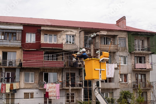 Fototapeta An electrician in a hard hat in a yellow aerial work platform repairs city lighting after broken wires on utility poles in Georgia, with a multi-story building in the background.