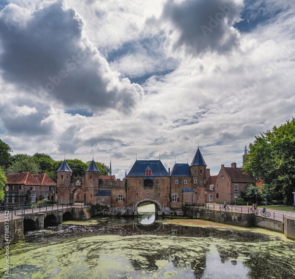 Fototapeta A picturesque medieval gate of the city of Amersfoort with pointed towers stands over a calm canal. The sky is filled with dramatic clouds, adding to the historic ambiance.