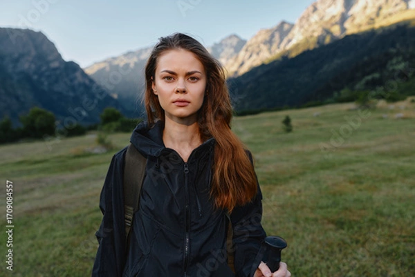 Fototapeta A focused young woman stands in a scenic mountain meadow, wearing a dark jacket and backpack. She gazes ahead with calm resolve, ready for an outdoor adventure.