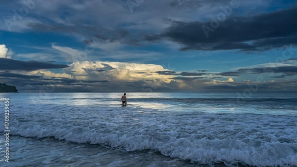 Fototapeta Early morning. The ocean waves are rolling towards the shore, foaming. Glare on the surface. The silhouette of a man knee-deep in water, walking into the sea. The clouds in the sky are highlighted  