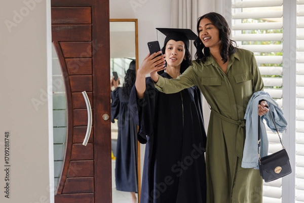 Fototapeta Mother and daughter taking selfie at home, celebrating graduation in cap and gown