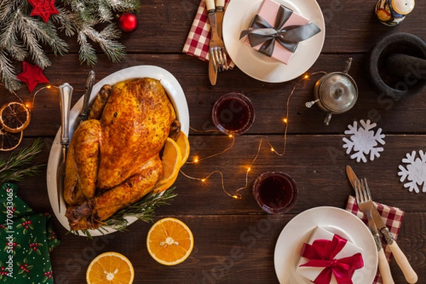 Fototapeta Christmas festive table is decorated with a garland. Baked chicken as a main holiday dish. Cone, dried oranges, plates, knife, fork furry spruce on a table. Top view.