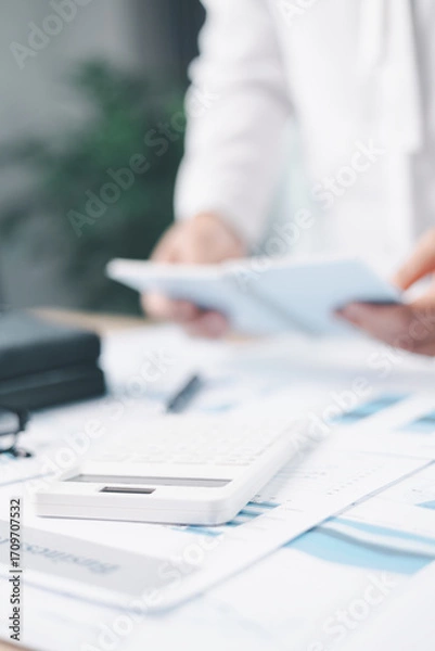 Fototapeta A businesswoman writes notes on clipboard while holding notebook at office desk with documents. Concept of business planning, accounting, financial management, productivity and corporate strategy.