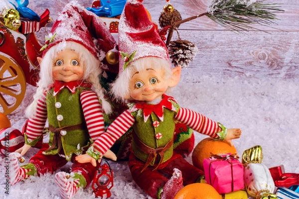 Fototapeta Funny christmas elves sits next to Christmas sock on light wooden background. Near tangerines and gifts.