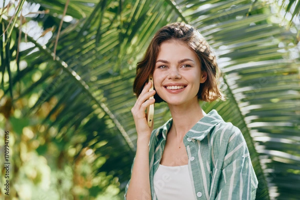 Fototapeta woman, portrait, smile, outdoors, phone call, casual clothes, green foliage, sunny day, cheerful mood, friendly gesture, natural light, candid moment, relaxed vibe, warm surroundings, bright