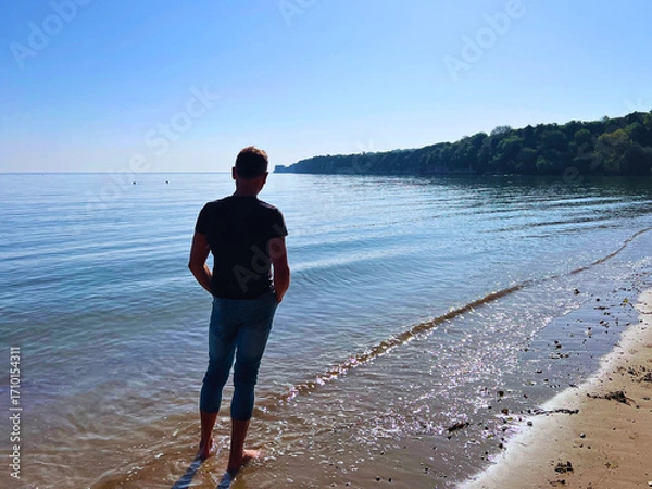 Fototapeta Slim man in jeans and top and bare feet on sandy Studland beach looking out to Old Harry Rock in Dorset UK.
