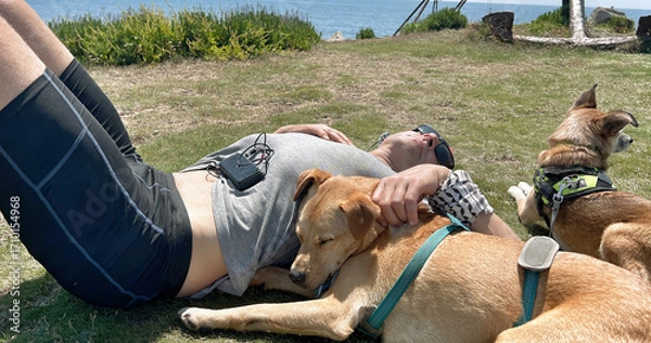 Fototapeta A fit slim man lying with dogs, sleeping & relaxing in a field on a sunny day in lycra shorts