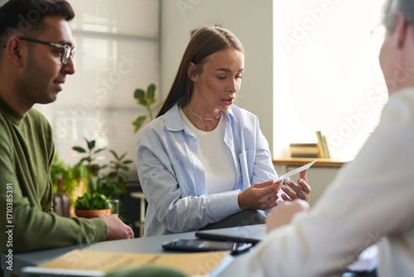 Fototapeta Woman holding paper while sitting with man and South Asian man, discussing psychological assessment in office setting