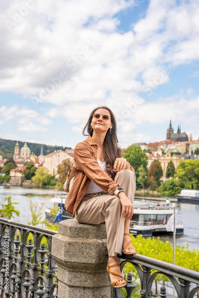 Fototapeta Young woman with a small backpack sitting on a stone railing with Prague Castle in the blurred background. Concept of solo travel, peaceful exploration and discovering historical European cities
