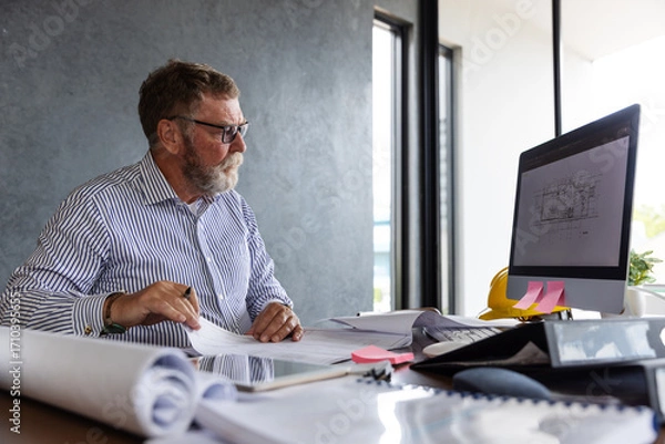 Fototapeta An older male architect or engineer reviews technical drawings at his desk.
