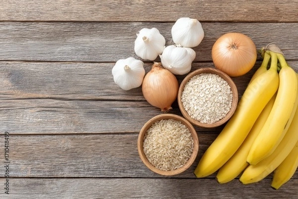 Fototapeta Fresh ingredients including bananas, onions, garlic, and bowls of rice and oats arranged on rustic wooden surface, showcasing healthy cooking essentials with copy space