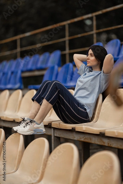 Fototapeta brunette girl is sitting comfortably in the empty stands of a stadium wearing a blue T shirt and casual pants. She appears relaxed enjoying the quiet atmosphere around her.