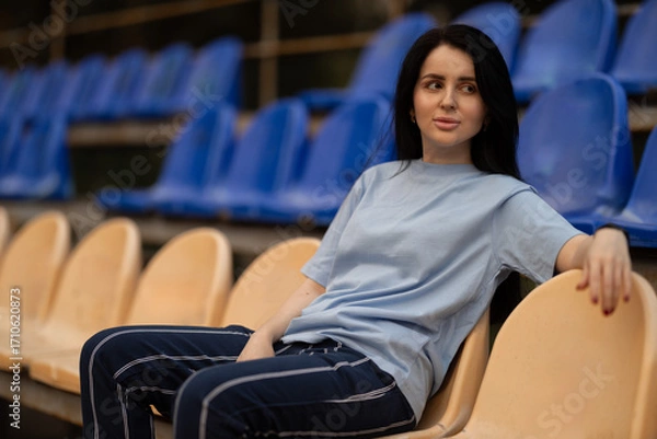 Fototapeta girl with beautiful brunette hair sits comfortably in the empty stands of a stadium wearing a blue t shirt. She gazes thoughtfully ahead while enjoying the peaceful atmosphere.