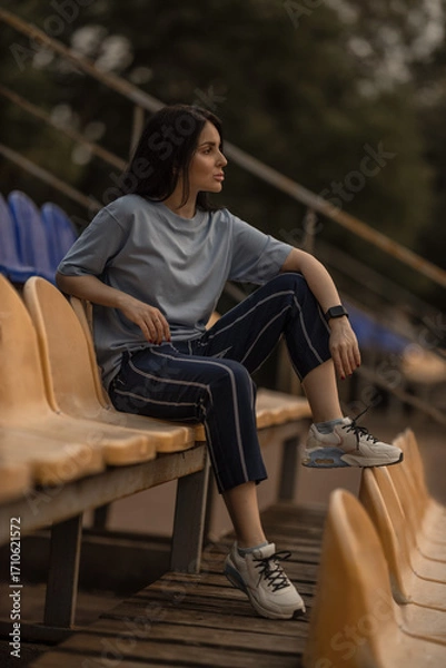 Fototapeta brunette girl relaxes alone in the empty stands of a stadium. She wears a blue t shirt and casual pants enjoying a peaceful moment in the sports venue.
