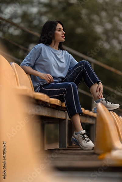 Fototapeta A brunette girl sits on the bleachers of a quiet stadium wearing a blue T shirt and casual pants. She looks thoughtfully into the distance as the sun sets creating a serene atmosphere.