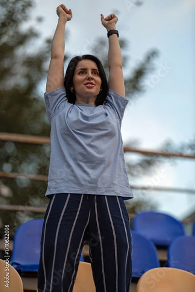 Fototapeta Brunette girl dressed in blue joyfully raises her hands on an empty podium celebrating a special moment in a public area. Excitement fills the air as she enjoys the day.