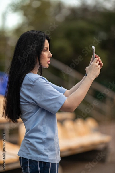 Fototapeta A brunette girl in a blue T shirt stands in the empty stands and focuses on filming a game on her phone. The surroundings are quiet and relaxed highlighting her concentration.