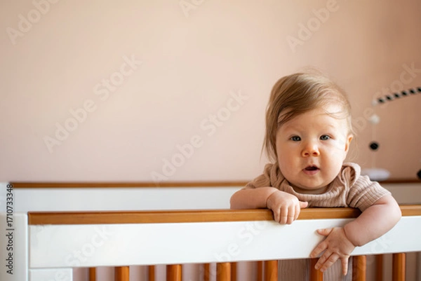Fototapeta Curious baby leaning over crib edge looking directly at camera in bright nursery setting, adorable toddler expression with natural light and cozy home family lifestyle concept