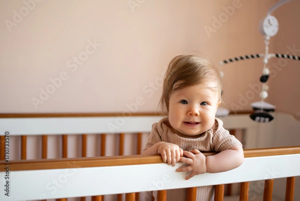 Fototapeta Adorable baby standing inside wooden crib smiling happily at camera in soft natural light, concept of childhood innocence, family home life, nursery comfort and baby lifestyle portrait