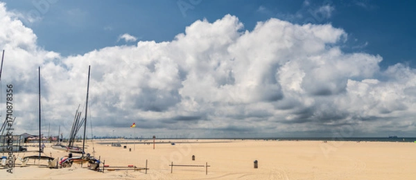 Fototapeta Catamarans and sailboats lie anchored on a vast, sandy beach under a sky filled with large, fluffy clouds. The expansive Kijkduin beach of The Hague stretches out to meet the horizon.