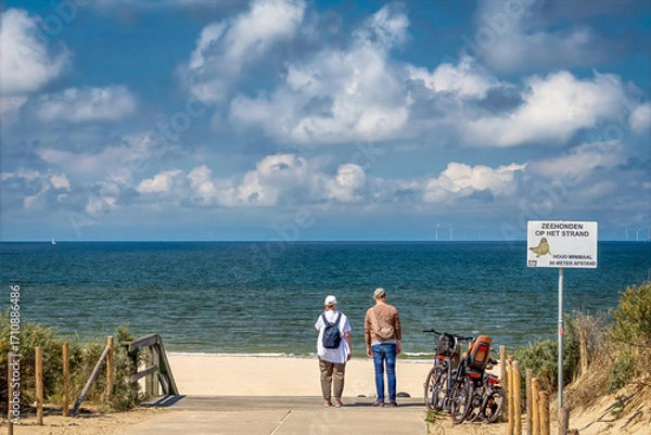 Fototapeta Two people stand at the end of a beach path, looking towards the ocean. Bicycles are parked nearby, and the sky is dotted with clouds.