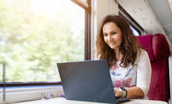 Fototapeta Young Caucasian woman smiling while working on laptop during train travel. Concept of digital lifestyle, remote work, and productive commuting on the go