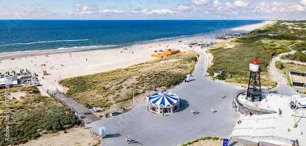 Fototapeta A panoramic view of a sandy beach of Kijkduin with a clear view of the sea. A lighthouse and a carousel are prominent near the shore, surrounded by pathways and greenery.