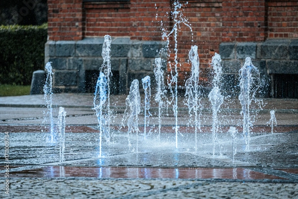Fototapeta Jets of water shoot up from a ground fountain in front of a brick wall, lit by glowing blue lights, creating a dynamic and refreshing urban scene.