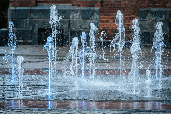Fototapeta Jets of water shoot up from a ground fountain in front of a brick wall, lit by glowing blue lights, creating a dynamic and refreshing urban scene.