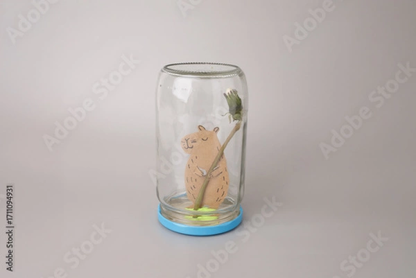 Fototapeta an upside-down glass jar resting on a blue base. Inside the jar, a handcrafted brown paper animal capybara holding delicate flower stem with a green bud tipped with white. creative process in action,