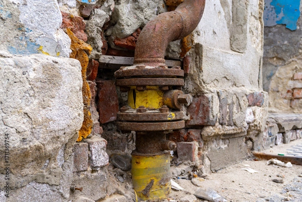 Fototapeta Old gas connection with yellow pipe and rusty valve, installed in damaged brick wall of deteriorating building structure.