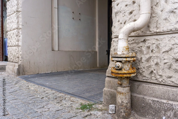 Fototapeta Gas connection with valve installed on tenement building façade, visible rust marks near entrance and cobblestone sidewalk