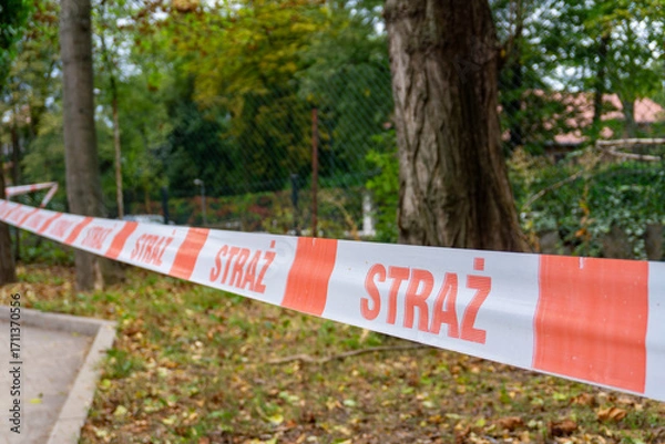 Fototapeta Warning tape with Polish inscription “STRAŻ” securing area near trees and sidewalk, marking restricted outdoor emergency scene.