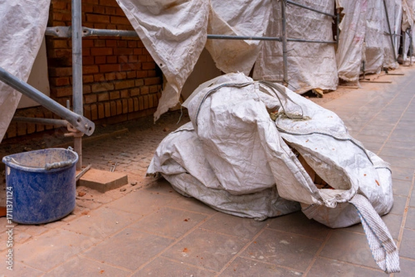 Fototapeta Large construction bag covered with tarp lying on renovation site near scaffolding and brick wall.